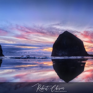 May include: A silhouette of Haystack Rock, a large sea stack, with two smaller sea stacks in the foreground, reflected in the water at sunset. The sky is a vibrant mix of pink, purple, and blue.
