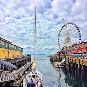 May include: A white sailboat docked in a harbor with a Ferris wheel in the background. The sailboat is in the foreground and the Ferris wheel is in the background. The sky is cloudy and the water is calm.