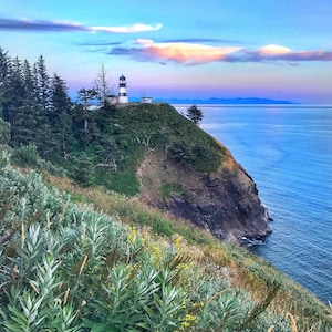 May include: A white and black lighthouse stands on a cliff overlooking a blue ocean. The sky is a gradient of blue, pink, and purple with white clouds. Green trees and bushes surround the lighthouse.