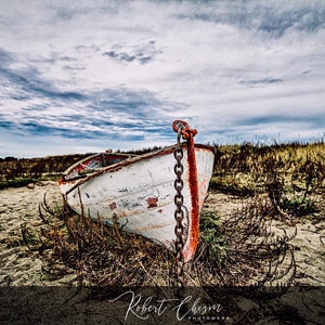 May include: A weathered white rowboat with red accents rests on a sandy beach, secured by a chain. The boat is surrounded by dry grass under a cloudy sky. The image has a vintage, rustic feel.