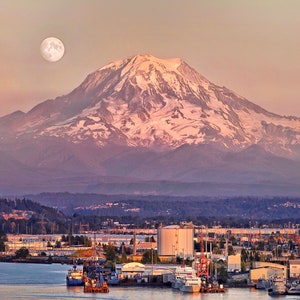 May include: A full moon hangs in the sky above a snow-capped mountain range, with a cityscape in the foreground. The city is located on a body of water, and there are several boats docked at the waterfront.