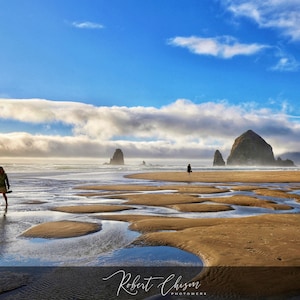 May include: A lone figure walks along a sandy beach with tide pools, towards Haystack Rock, a large rock formation in the distance. The sky is a bright blue with white clouds.