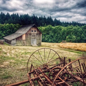 May include: A weathered wooden barn with a rusty farm implement in the foreground. The barn has a wooden door and windows. The sky is cloudy and gray. The implement is a large, rusty wheel with spokes.