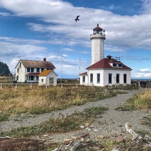 May include: A white lighthouse with a red roof stands on a grassy hill overlooking the ocean. A smaller white building with a red roof is to the left of the lighthouse. The sky is blue with white clouds.