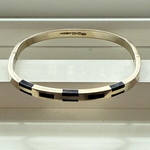May include: A gold-toned hinged bangle bracelet with a geometric design. The bracelet features alternating black and gold rectangular inlays. The text "MERIT CR-18GD" is stamped on the inside of the bracelet.