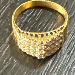 May include: A gold ring featuring a wide, elevated band set with multiple small, clear, faceted stones arranged in a cluster across the top. The metal has a polished yellow finish, and the design includes a decorative, scalloped gallery setting.