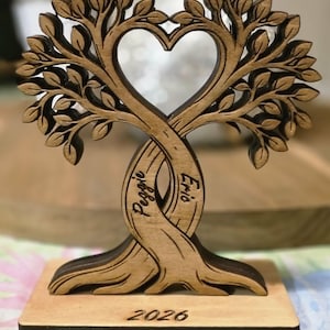 May include: A wooden tree sculpture with a heart-shaped opening in the center. The intertwined trunks feature the names "Peggie" and "Eric". The base displays the year "2026". The sculpture is made of light brown wood.