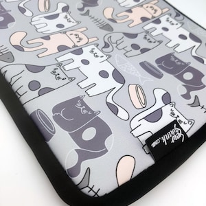 Puede incluir: Una funda gris para portátil con un patrón repetido de gatos de dibujos animados en varias poses, con acentos blancos, grises y rosas. El diseño incluye espinas de pescado y cuencos de comida. La funda tiene un borde negro y una etiqueta que dice "sewfrank.com".