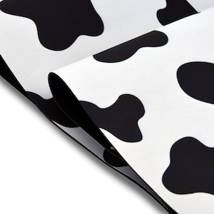 May include: Close-up of a white fabric with large black cow spots. The fabric is folded over, revealing the underside. The pattern is repeated across the surface, creating a playful and bold design.
