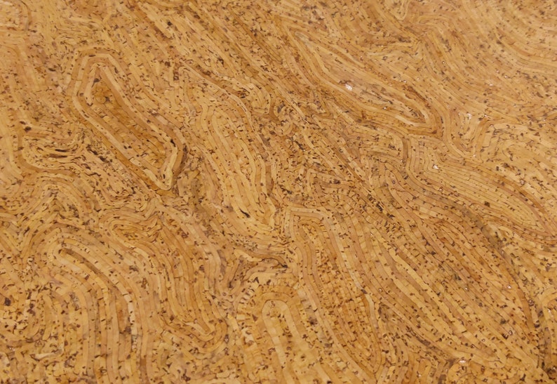 May include: Close-up of a textured, light brown cork surface. The cork has a layered, wavy pattern with darker specks throughout. The design resembles a topographical map or the rings of a tree.