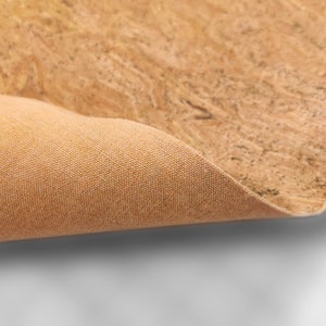 May include: Close-up of a sheet of cork material with a natural, textured surface. The cork has a light brown color with darker brown and black specks. The underside of the cork is a tan fabric.