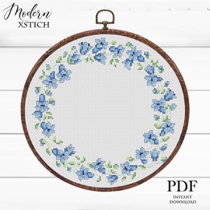 May include: A cross-stitch design in a wooden hoop, showcasing a wreath of blue flowers and green leaves. The text "Modern XSTICH" is at the top left, and "PDF INSTANT DOWNLOAD" is at the bottom right.