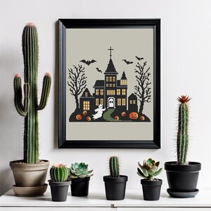 May include: A black and white cross-stitch pattern of a spooky Halloween house with a ghost, bats, and pumpkins. The house has a pointed roof and windows with glowing lights. The pattern is framed in a black frame and displayed on a white wall.