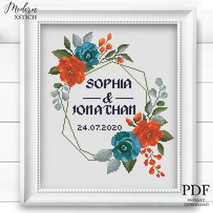 May include: Framed cross-stitch artwork featuring a floral wreath in shades of orange, teal, and green. The names "SOPHIA & JONATHAN" are centered within a geometric shape, with the date "24.07.2020" below. The frame is white.