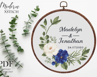 May include: Cross stitch pattern featuring a floral wreath with white and blue flowers, green leaves, and the text "Madelyn & Jonathan 24.07.2020".