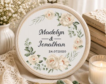 Puede incluir: Un bastidor de bordado circular de madera que contiene un diseño de punto de cruz de rosas color crema y follaje verde. El texto dice Madelyn & Jonathan con la fecha 24.07.2020 centrada dentro de la corona floral sobre tela blanca.