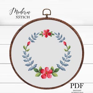 Könnte beinhalten: Ein Kreuzstich-Stickrahmen mit einem floralen Kranz-Design. Der Kranz umfasst rote Blumen, blaue Blätter und grüne Akzente. Der Text "Modern XSTICH" befindet sich oben und "PDF INSTANT DOWNLOAD" unten.