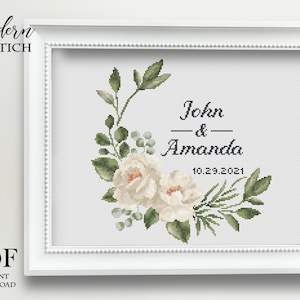 May include: Cross stitch pattern featuring a white floral wreath with green leaves and the text "John & Amanda 10.29.2021".
