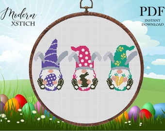 May include: Three gnomes with Easter-themed decorations. The gnome on the left is wearing a purple hat with white polka dots and holding a purple egg with white polka dots. The gnome in the middle is wearing a pink hat with white polka dots and holding a white bunny. The gnome on the right is wearing a green hat with white flowers and holding a carrot.