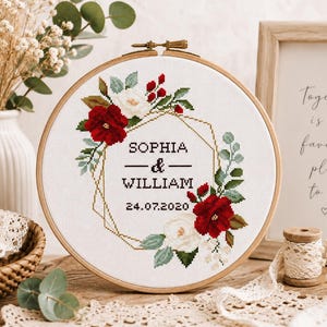 Könnte beinhalten: Ein runder Holzstickrahmen mit weißem Stoff, der einen im Kreuzstich gestickten Blumenkranz in Rot und Grün zeigt. In der Mitte befindet sich der Text SOPHIA & WILLIAM sowie das Datum 24.07.2020, umrahmt von einem geometrischen Goldmuster.