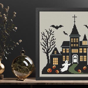 May include: A black and white cross-stitch pattern of a spooky haunted house with a ghost, bats, and pumpkins. The house has lit windows and a front door. The background is a light grey.