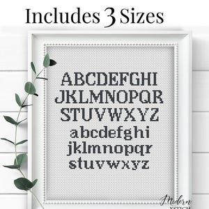 May include: A framed cross-stitch alphabet sampler in a white frame. The sampler features uppercase and lowercase letters in a dark grey colour. The text "Includes 3 Sizes" is at the top. The Modern XStitch logo is at the bottom.