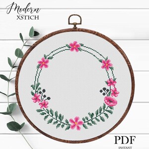 Puede incluir: Un diseño de corona floral de punto de cruz en un bastidor de bordado de madera. La corona presenta flores rosas y hojas verdes, con el texto "Modern XSTICH" en la parte superior y "PDF Instant Download" en la inferior. El bastidor tiene unos 20 cm de diámetro.