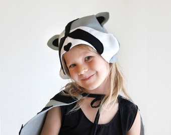 May include: A child in a raccoon costume, featuring a hat and cape. The hat is grey, white, and black, with a raccoon mask design and ears. The cape is grey with black details. The child is wearing a black top.