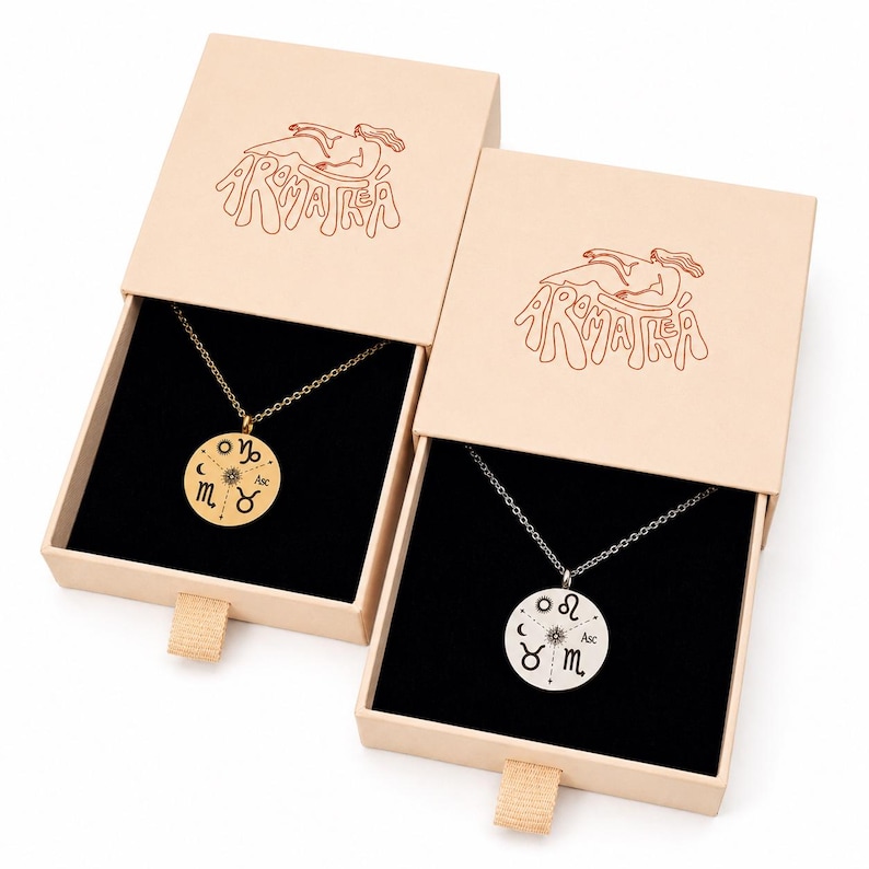 May include: Two necklaces with round pendants featuring engraved zodiac symbols and a central sun icon, presented in beige sliding gift boxes. Each box lid displays a line drawing of a reclining figure and the text AROMATEA in red ink.