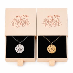 May include: Two round pendant necklaces, one silver-toned and one gold-toned, each featuring engraved zodiac symbols and a central sun icon. The necklaces are presented in individual light beige sliding boxes with the text AROMATEA printed.