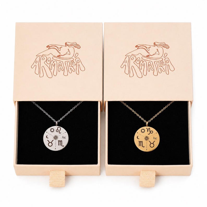 May include: Two round pendant necklaces, one silver-toned and one gold-toned, each featuring engraved zodiac symbols and a central sun icon. The necklaces are presented in individual light beige sliding boxes with the text AROMATEA printed.
