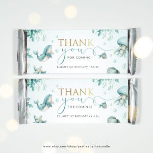 May include: A set of two blue and white chocolate bar wrappers with a thank you message for a first birthday party. The wrappers feature a whale, jellyfish, starfish, and seashells. The text on the wrappers reads "Thank you for coming! Elijah's 1st Birthday - 8.8.26".