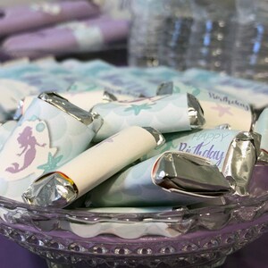 Puede incluir: Un bol de vidrio lleno de peque&ntilde;as barras de chocolate envueltas en plata. Los envoltorios est&aacute;n decorados con una sirena, conchas marinas y el texto "Feliz cumplea&ntilde;os".