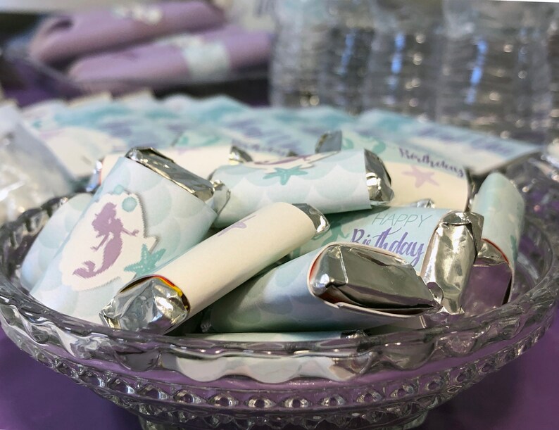 Puede incluir: Un bol de vidrio lleno de peque&ntilde;as barras de chocolate envueltas en plata. Los envoltorios est&aacute;n decorados con una sirena, conchas marinas y el texto "Feliz cumplea&ntilde;os".