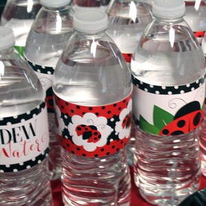 May include: Clear plastic water bottles with white caps. The bottles have labels with ladybug designs and the words "Dew Water" and "Love Bug" in red script. The labels feature red and black polka dots.