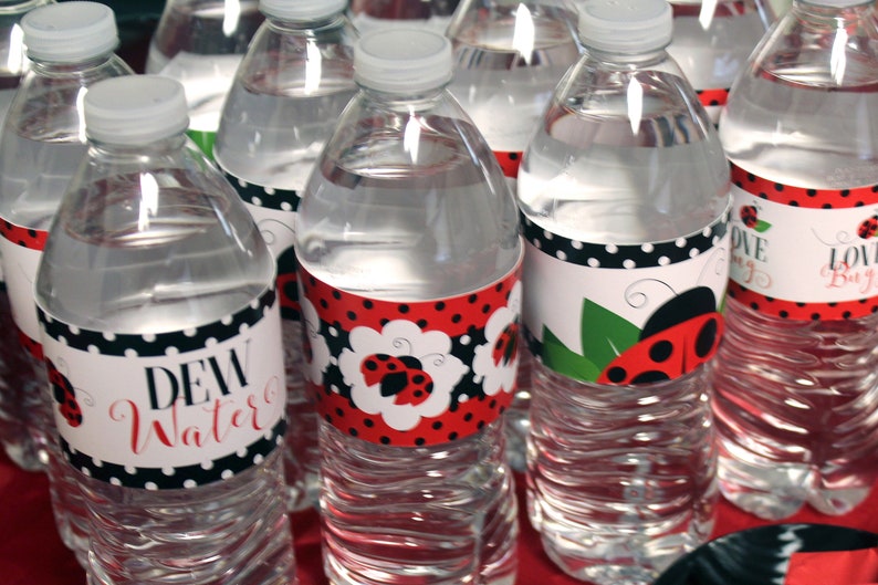 May include: Clear plastic water bottles with white caps. The bottles have labels with ladybug designs and the words "Dew Water" and "Love Bug" in red script. The labels feature red and black polka dots.