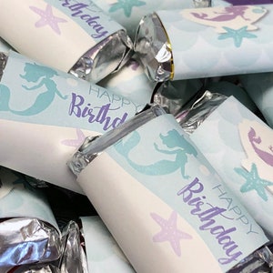 Puede incluir: Primer plano de un mont&oacute;n de peque&ntilde;as barras de chocolate envueltas en papel de aluminio plateado y decoradas con envoltorios de papel con tem&aacute;tica de sirenas. Los envoltorios son de color azul claro con detalles morados y presentan el texto "Feliz cumplea&ntilde;os" y una ilustraci&oacute;n de sirena.