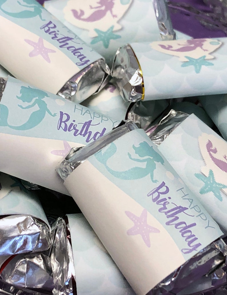 Puede incluir: Primer plano de un mont&oacute;n de peque&ntilde;as barras de chocolate envueltas en papel de aluminio plateado y decoradas con envoltorios de papel con tem&aacute;tica de sirenas. Los envoltorios son de color azul claro con detalles morados y presentan el texto "Feliz cumplea&ntilde;os" y una ilustraci&oacute;n de sirena.