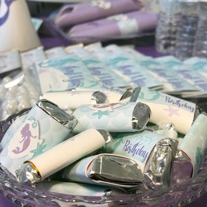 Puede incluir: Un taz&oacute;n de vidrio transparente lleno de barras de chocolate envueltas individualmente. Los envoltorios est&aacute;n decorados con un tema de sirena y el texto "Feliz cumplea&ntilde;os".