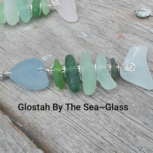 Puede incluir: Un collar de pez de vidrio de mar con un alambre de plata y una variedad de piezas de vidrio de mar verde y azul. El collar est&aacute; sobre una superficie de madera y el texto "Glostah By The Sea~Glass" es visible.