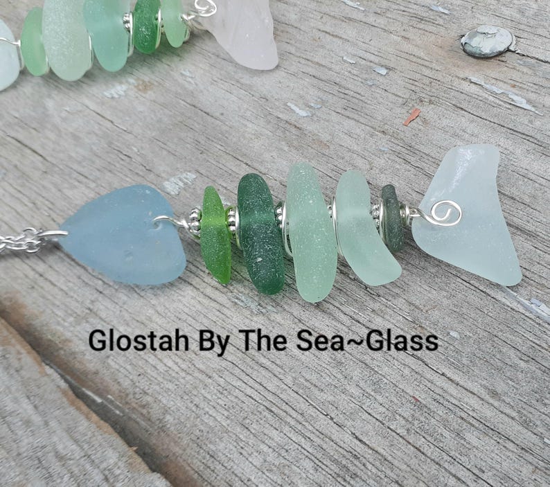 Puede incluir: Un collar de pez de vidrio de mar con un alambre de plata y una variedad de piezas de vidrio de mar verde y azul. El collar est&aacute; sobre una superficie de madera y el texto "Glostah By The Sea~Glass" es visible.