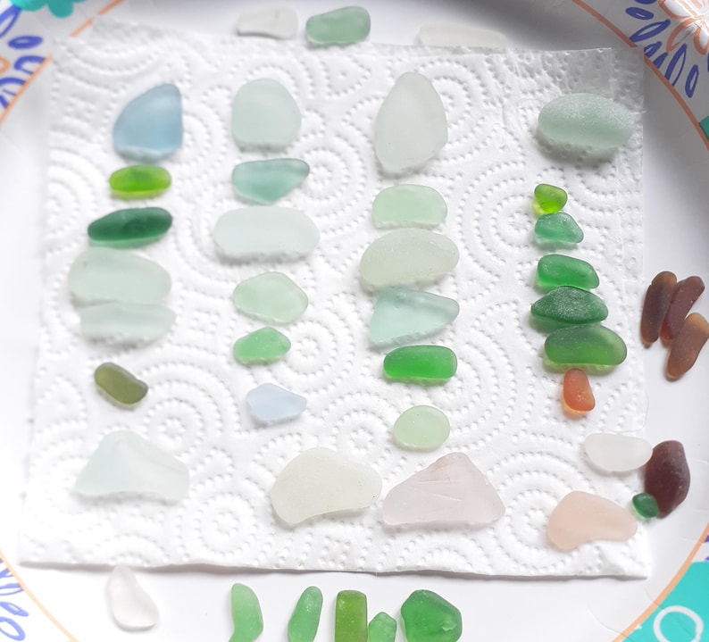 Puede incluir: Una colecci&oacute;n de piezas de vidrio de mar en varios tonos de verde, azul y blanco, dispuestas sobre una toalla de papel blanca. El vidrio de mar est&aacute; ordenado por color y tama&ntilde;o, con algunas piezas apiladas unas sobre otras.
