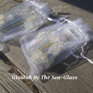 May include: Several small, white, translucent, leaf-shaped lights are inside sheer white drawstring bags. The bags are on a weathered wooden surface. The text "Glostah By The Sea~Glass" is visible at the bottom of the image.