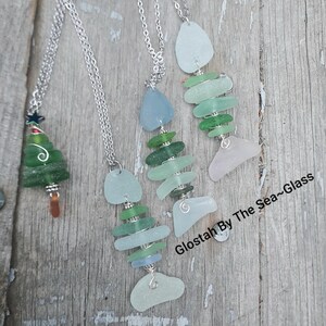 Puede incluir: Cuatro collares con colgantes de vidrio de mar en forma de &aacute;rbol de Navidad y pez. Los colgantes en forma de pez est&aacute;n hechos de varios tonos de vidrio de mar verde y azul. Los collares est&aacute;n sobre una superficie de madera con el texto "Glostah By The Sea-Glass" en la esquina inferior derecha.