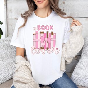 Peut inclure: T-shirt blanc avec un graphique rose et marron de livres empilés avec un nœud rose et le texte "*BOOK lover*"