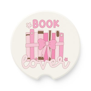 May include: A white ceramic coaster with a pink and brown design. The coaster features a stack of books tied with a pink bow and the text "*BOOK lover*" with small pink flowers.