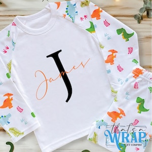 Può includere: Set pigiama bianco con maniche lunghe e pantaloni abbinati. Le maniche e i pantaloni presentano un motivo di dinosauri colorati. La maglietta ha una grande lettera nera "J" con il nome "James" in arancione. Il testo "that's a WRAP" è visibile.