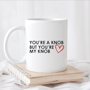 May include: White ceramic mug with the text "YOU'RE A KNOB BUT YOU'RE MY KNOB" in black, and a red heart. The mug is resting on a small stack of books.
