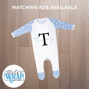 Puede incluir: Mono de bebé blanco con mangas y pies con estampado de nubes azules. El mono presenta una gran letra negra "T" y el nombre "Tommy" en escritura azul claro. El texto "MATCHING PJ'S AVAILABLE" está en la parte superior.