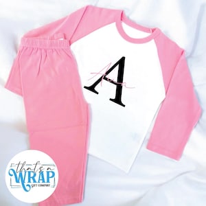 Puede incluir: Un conjunto de pijama rosa y blanco. La parte superior de manga larga tiene mangas rosas y un cuerpo blanco con una gran letra negra "A" y el nombre "Ava" en cursiva. Los pantalones de pijama a juego son rosas. La imagen también incluye el texto "that's a WRAP gift company".